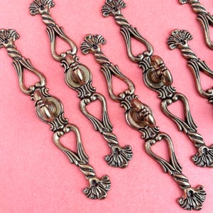 May include: Antique brass-toned cabinet pulls with ornate detailing. Each pull features a central circular design with a decorative handle and a twisted, textured bar. The pulls are arranged diagonally against a pink background.