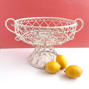 May include: A white, ornate wire fruit basket with a pedestal base and two handles. The basket is filled with a few yellow lemons. The background is a solid pink color.