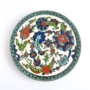 May include: Round ceramic coaster with a floral design. The coaster features a white background with a vibrant pattern of red, blue, and green flowers and leaves. A green geometric border frames the design.