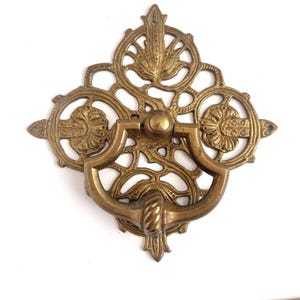 May include: A brass door knocker featuring an intricate, openwork backplate with a symmetrical, scrolling design. The central element is a U-shaped, hinged handle with a textured, rope-like detail at the bottom and a smooth, rounded top knob.