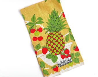May include: A folded yellow linen towel with a vibrant fruit pattern. The design features a large pineapple, red strawberries, and cherries with green leaves. The towel has a white border and a label that reads "ALL PURE LINEN".