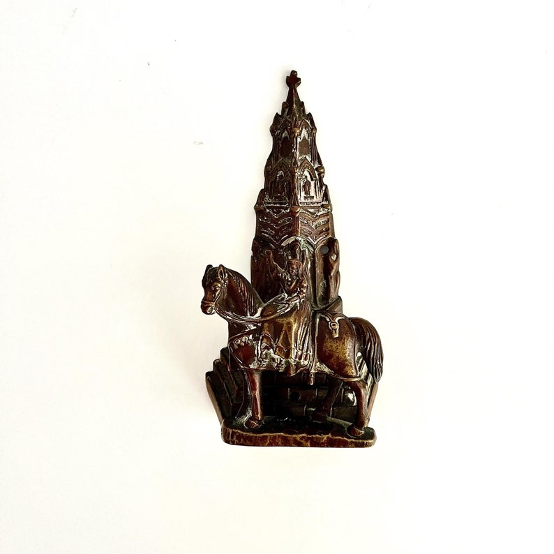 May include: A bronze door knocker in the shape of a knight riding a horse in front of a church. The church has a pointed roof and a cross on top.
