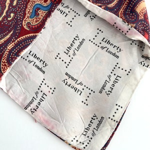 May include: A white fabric with the text "Liberty of London" repeated in a circular pattern. The fabric is folded over a red and blue paisley patterned fabric.