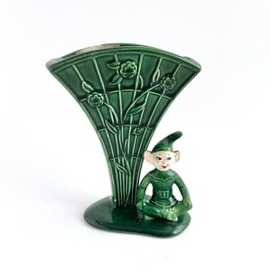 May include: A dark green ceramic vase featuring a fan-shaped body with embossed floral trellis patterns. Attached to the base is a small, seated elf figurine with a pointed hat, painted with a pale face, dark eyes, and red lips, all in a glossy finish.