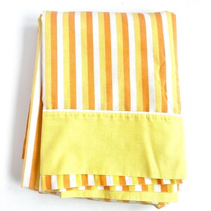 May include: A folded yellow and orange striped fabric with a solid yellow border. The fabric is likely a bed sheet or a curtain.