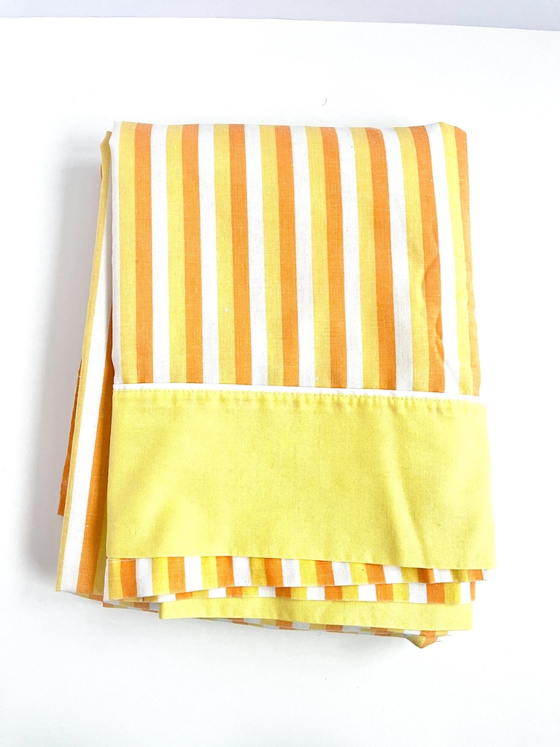 May include: A folded yellow and orange striped fabric with a solid yellow border. The fabric is likely a bed sheet or a curtain.