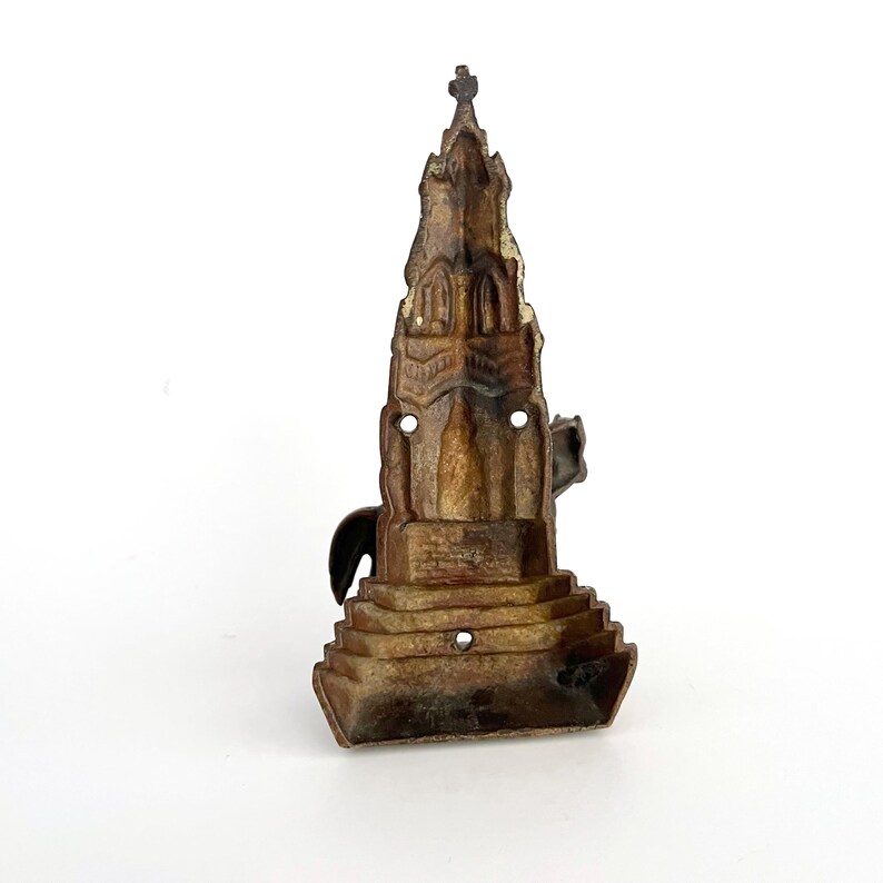 May include: A cast iron wall mounted doorstop in the shape of a church with a pointed roof and a cross on top. The doorstop is painted in a brown color and has a worn, aged look.