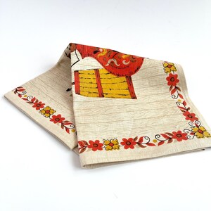 May include: Folded vintage beige linen napkin with a colorful print. The design features a red and yellow illustration and a floral border with red and yellow flowers. The napkin has a wood grain pattern.