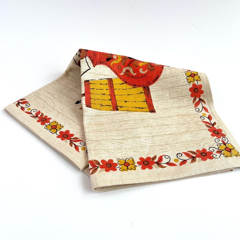 May include: Folded vintage beige linen napkin with a colorful print. The design features a red and yellow illustration and a floral border with red and yellow flowers. The napkin has a wood grain pattern.