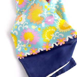 May include: A colorful silk scarf with a floral pattern. The scarf features a light blue background with large flowers in yellow, pink, and orange. The bottom portion is a solid navy blue with a white scalloped edge.