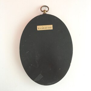 May include: Black oval-shaped picture frame with a brass hook on top. The back of the frame is black and has a label that reads "16 BLACK-IVORY WOOD".