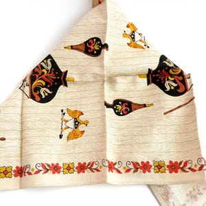 May include: Cream-colored vintage kitchen towel with a repeating pattern of black, brown, yellow, and red decorative elements. The design includes stylized bottles, eagles, and floral borders, evoking a retro aesthetic.