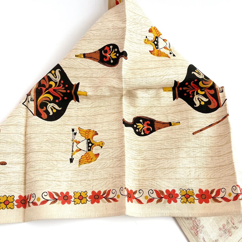 May include: Cream-colored vintage kitchen towel with a repeating pattern of black, brown, yellow, and red decorative elements. The design includes stylized bottles, eagles, and floral borders, evoking a retro aesthetic.