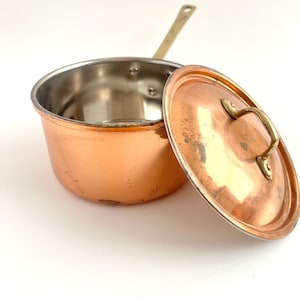 May include: A round copper saucepan with a polished silver-toned interior and a long metal handle. The matching copper lid features a curved brass handle and rests beside the pan, showing a warm metallic finish and signs of light wear.