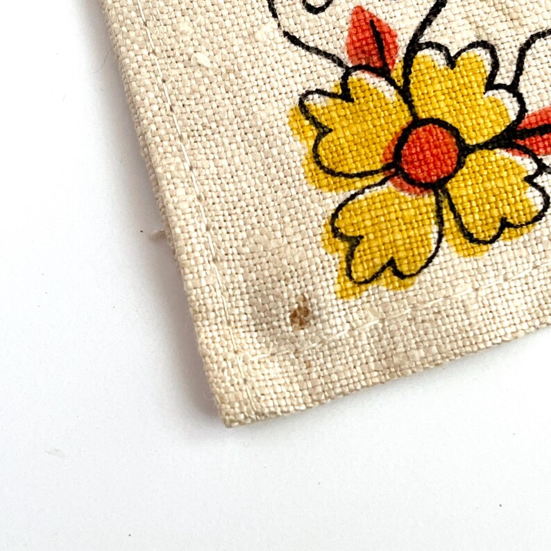 May include: Close-up of a section of a beige linen fabric with a floral design. The flower has yellow petals, a red center, and black outlines. The fabric has a visible weave and a few small brown spots.