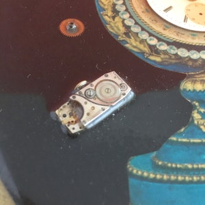 May include: A close-up of a silver watch movement with exposed gears and a small, round dial. The movement is lying on a dark surface.