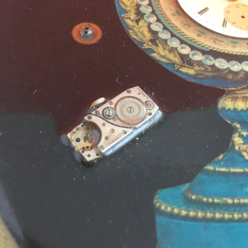 May include: A close-up of a silver watch movement with exposed gears and a small, round dial. The movement is lying on a dark surface.
