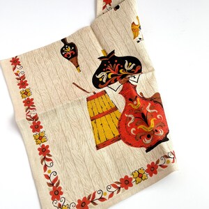 May include: A vintage cream-colored kitchen towel with a repeating pattern of stylized figures and objects in red, yellow, and black. The towel has a floral border in red and yellow. The design includes a barrel, a jug, and a figure wearing a hat.
