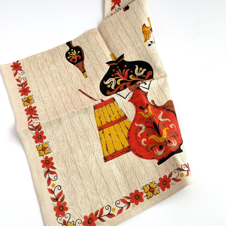 May include: A vintage cream-colored kitchen towel with a repeating pattern of stylized figures and objects in red, yellow, and black. The towel has a floral border in red and yellow. The design includes a barrel, a jug, and a figure wearing a hat.