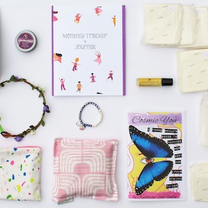 May include: A flat lay of a self-care package with a variety of items, including a journal, a book, a flower crown, a pouch, a tea bag, a bath bomb, a bracelet, a bottle of essential oil, and a set of reusable pads.