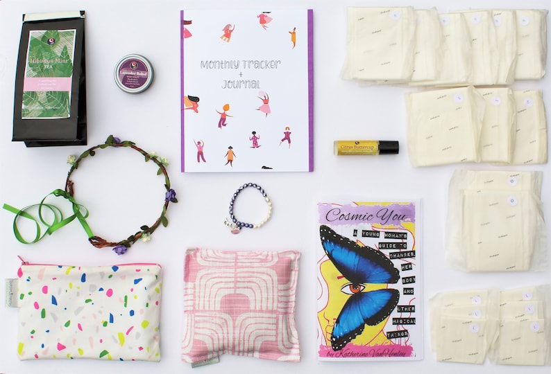 May include: A flat lay of a self-care package with a variety of items, including a journal, a book, a flower crown, a pouch, a tea bag, a bath bomb, a bracelet, a bottle of essential oil, and a set of reusable pads.