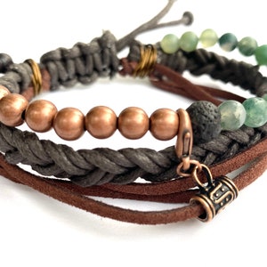 Puede incluir: Un brazalete de múltiples hebras con cordón de cuero marrón, cordón trenzado gris, cuentas de cobre y piedras de ágata verde. El brazalete tiene un pequeño dije de metal con un diseño.