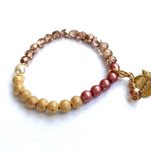 Op de afbeelding: Een gouden, bruine en witte kralen armband met een gouden bedel met de tekst "elene beads" en een stervormig ontwerp.