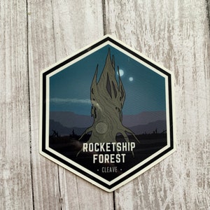 May include: A hexagonal sticker with a dark blue background and a stylized illustration of a tree with a circular pattern on its trunk. The text "ROCKETSHIP FOREST" is printed in white below the tree, with "CLEAVE" in smaller white text below that.