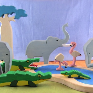 May include: A wooden toy set featuring a blue baobab tree, two adult elephants, two baby elephants, two pink flamingos, and two green crocodiles. The set includes a blue pond with a wooden base.