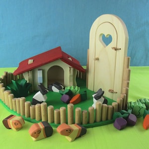 May include: A wooden toy farm set with a red-roofed house, a heart-shaped door, a picket fence, and wooden rabbits and guinea pigs. The set includes wooden carrots, beetroot, and bushes. The background is light blue and green.