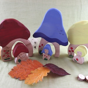 May include: A set of four wooden mushroom toys painted in red, blue, yellow, and natural wood. Each mushroom has a small figure of a person with a painted face and flowers. The mushrooms are arranged on a light brown surface with a few autumn leaves.