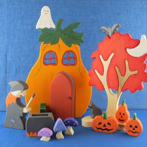 Könnte beinhalten: Ein hölzernes Halloween-Spielset mit einem Kürbishaus, einer Hexe, einem Kessel, einem Baum mit Geistern und Kürbissen. Das Kürbishaus hat eine rote Tür und Fenster. Die Hexe hält einen Stock. Der Baum ist rot mit einem braunen Stamm. Die Kürbisse sind orange mit schwarzen Gesichtern.