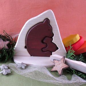 May include: Wooden toy puzzle featuring a brown bear and cub nestled in a cave, with a stack of colourful wooden rings and a star-shaped piece.