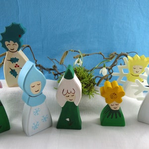 May include: Seven wooden figurines painted in various colors, representing different flowers. The figurines are arranged on a white surface with a branch of a tree behind them. The flowers include a pink snowdrop, a blue snowdrop, a white snowdrop, a yellow daffodil, a green snowdrop, a green holly, and a white snowflake.