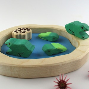Könnte beinhalten: Holzspielzeugteich mit einem grünen Frosch, der am Rand sitzt, und drei grünen Fröschen, die im Wasser schwimmen. Der Teich ist aus hellem Holz und hat eine blaue Wasseroberfläche.