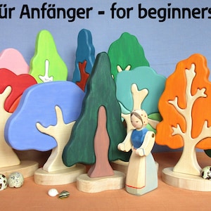 Könnte beinhalten: Ein Set aus bunten Holzspielzeugbäumen mit einer kleinen Holzfigur, die davor steht. Die Bäume sind alle in verschiedenen Formen und Größen und in leuchtenden Farben bemalt. Die Figur trägt ein blaues Kleid und eine weiße Schürze. Der Text "für Anfänger - for beginners" ist oben im Bild gedruckt.