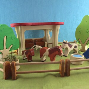 May include: Wooden toy farm set with a barn, fence, trees, and toy horses. The horses are brown and white. The barn has a red roof and the trees are green. A blue and yellow flag is also included.