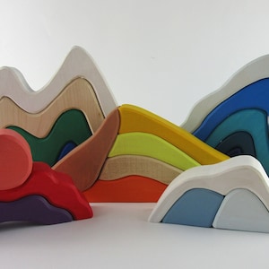 Puede incluir: Un conjunto de juguetes de madera de colores que incluye una ola, montañas y un sol. La ola es azul, verde y blanca. Las montañas son blancas, marrones, verdes, amarillas y naranjas. El sol es rojo.