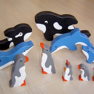 May include: Wooden toy set featuring a black and white orca, blue dolphins, and gray penguins with orange beaks and feet. The aquatic animal figures are painted with smooth, clean lines, perfect for imaginative play.