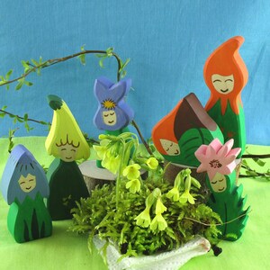 Pode incluir: Figuras de madeira de flores com rostos, pintadas em tons de azul, verde, amarelo, laranja e rosa. As figuras estão dispostas sobre um leito de musgo e rodeadas de pequenas flores amarelas.