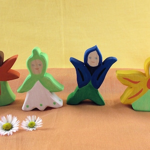 May include: A set of seven wooden figurines painted to resemble different flowers. The figurines are all wearing different colored clothing and have different facial expressions. The figurines are all standing on a light brown surface.