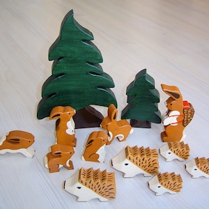 May include: A set of wooden animal figurines, including a rabbit carrying a basket, a hedgehog, and a squirrel. The figurines are painted in brown, white, and green colours. The set also includes two green wooden trees.