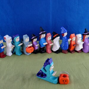 Może przedstawiać: Rząd drewnianych figurek Halloweenowych ubranych w kostiumy, w tym czarownica, duch, wampir i dynia. Figurki trzymają torby na cukierki i stoją na zielonej powierzchni.