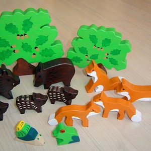 May include: A set of wooden animal figurines, including a boar, piglets, foxes, and a frog. The figurines are painted in realistic colors and are arranged around two green trees with brown trunks.