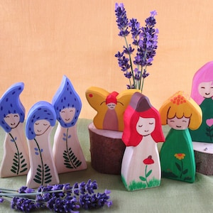 May include: A set of wooden figurines painted in various colors, including blue, red, yellow, green, and pink. The figurines depict people with different hairstyles and clothing. The figurines are arranged on a wooden surface with a bouquet of lavender flowers in the background.