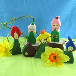 Op de afbeelding: Een set van zeven houten figuurtjes die gestileerde bloemen voorstellen. De bloemen zijn in verschillende kleuren geschilderd, waaronder rood, roze, geel, blauw en groen. De figuurtjes zijn gerangschikt op een groene ondergrond met twee gele narcissen.