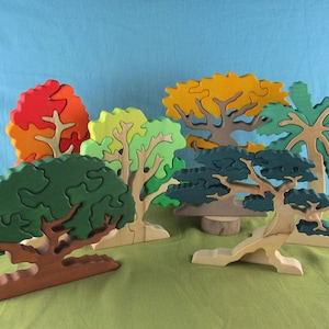 May include: A collection of colorful wooden tree puzzles in various shapes and sizes. The trees feature shades of green, red, yellow, and blue, with detailed trunk and branch designs. The puzzles are displayed on a green surface against a blue backdrop.