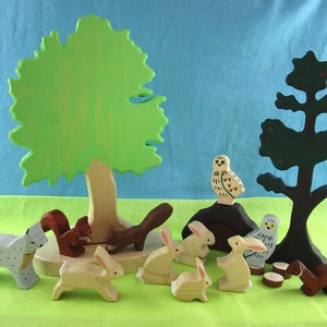 May include: A set of wooden animal figurines, including a squirrel, a lynx, a rabbit, an owl, and a weasel, arranged around a green tree and a black tree. The figurines are painted in realistic colors and have detailed features.