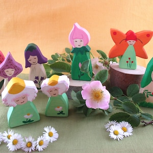 May include: A set of wooden figurines painted in bright colors, depicting fairies and flowers. The fairies are wearing green, pink, and purple dresses and have painted faces. The flowers are painted in shades of pink, red, and yellow. The figurines are arranged on a green surface with small white flowers.