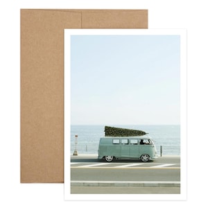 Può includere: Fotografia di un furgone vintage verde chiaro che guida su una strada costiera con un albero di Natale sul tetto. Il furgone è di fronte al mare sotto un cielo azzurro. L'immagine è su sfondo bianco con una busta marrone.